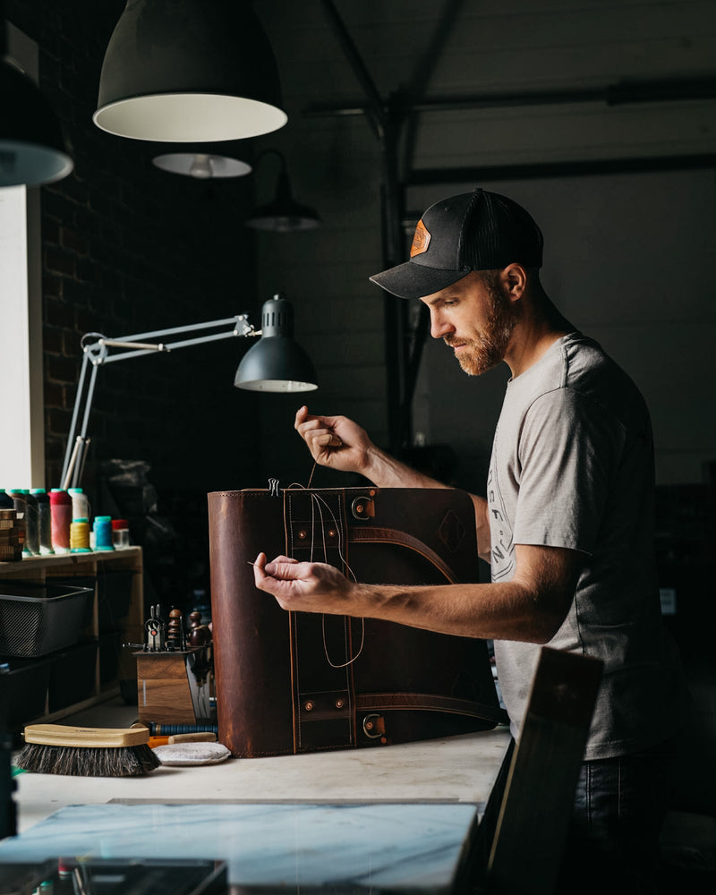 A man in a workshop wearing a black baseball cap and gray t-shirt, holding and inspecting a dark brown leather bag with leather straps and buckles.
