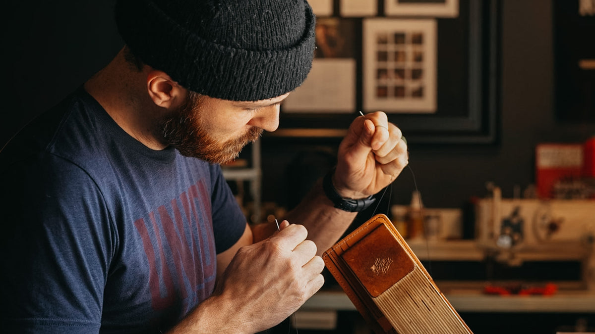 A man wearing a black knit beanie and a navy blue graphic t-shirt is shown in a workshop setting, holding a small wooden object in his hands. The background features warm, dimly lit shelves and framed pictures.