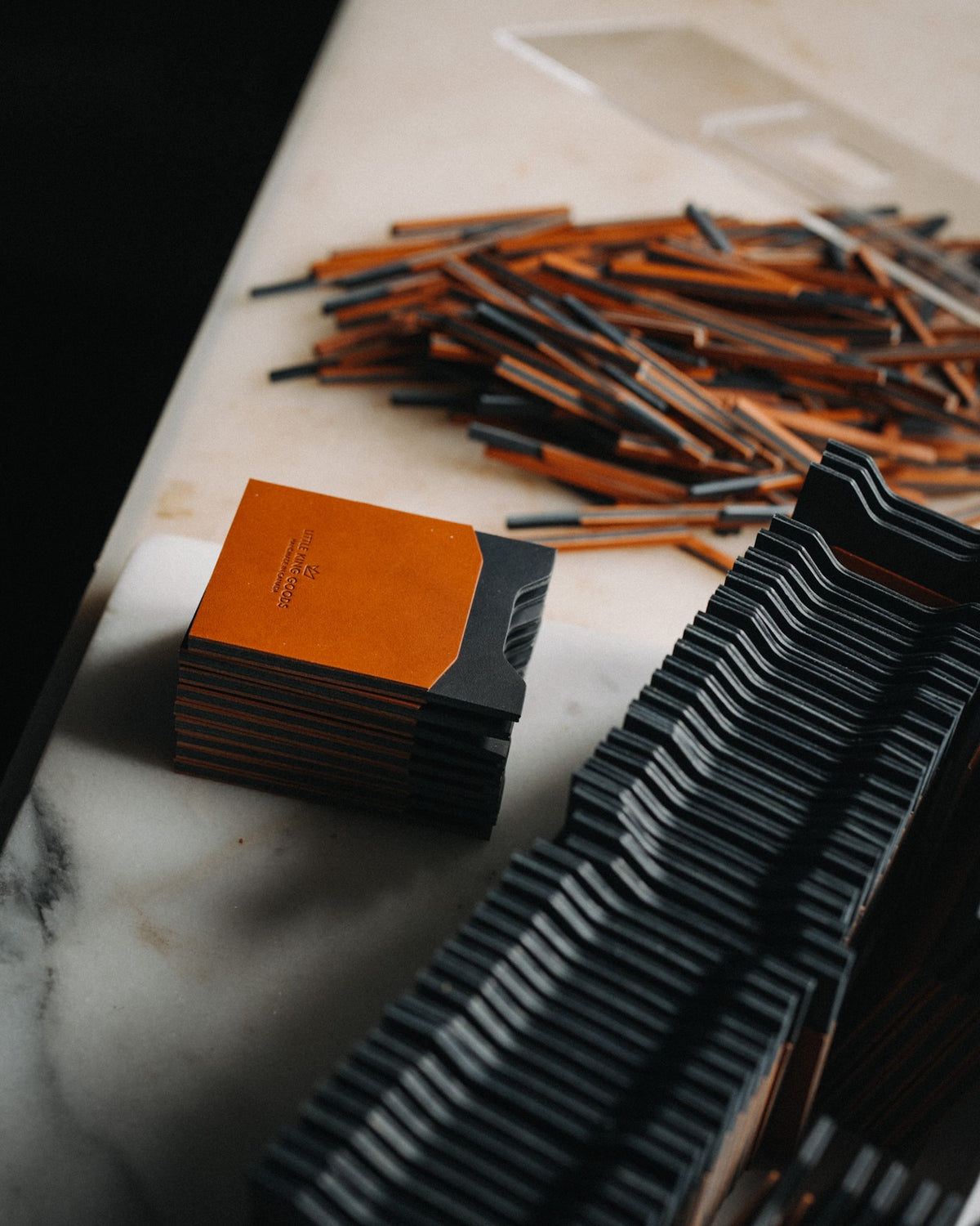 A stack of slim rectangular cards or cases with a matte orange top and black sides sits on a white marble surface, with many matching pieces scattered and stacked in the background. No person or animal is present, and the scene is styled as a clean product shot rather than a real-use environment, so subject_type is Product/Packaging.